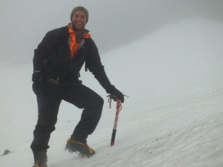 A man poses with ice axe and crampons in a white out on a mountain.
